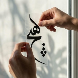 May include: A black vinyl decal with Arabic script is being applied to a window. The decal is on a clear backing, and the hands are peeling the backing away. The script is a stylized, flowing design.