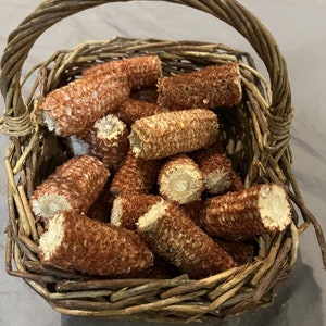 May include: A woven wicker basket filled with multiple dried, reddish-brown corn cobs. The basket features a sturdy arched handle and a square base, containing several stripped corn cobs with visible textures and light-colored, circular ends.