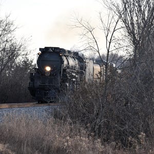 May include: A large, black steam locomotive, number 3401, travels along a railroad track. The train is surrounded by trees and brush, with a bright headlight illuminating the way. Smoke billows from the engine, creating a dramatic scene.