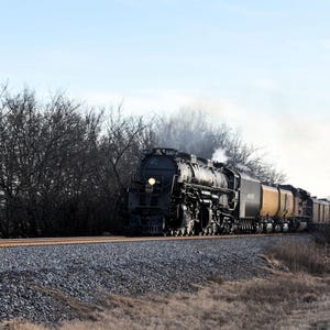 May include: A large black steam train with yellow and black cars travels on a gravel track. The train is moving left to right, with steam rising from the engine. Bare trees line the tracks under a blue sky.