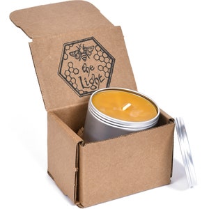 Puede incluir: Una vela de miel en una lata de plata con una caja de cartón. La caja está abierta y la vela está dentro. La caja tiene un logotipo que dice "The Light" con un diseño de abeja y panal.