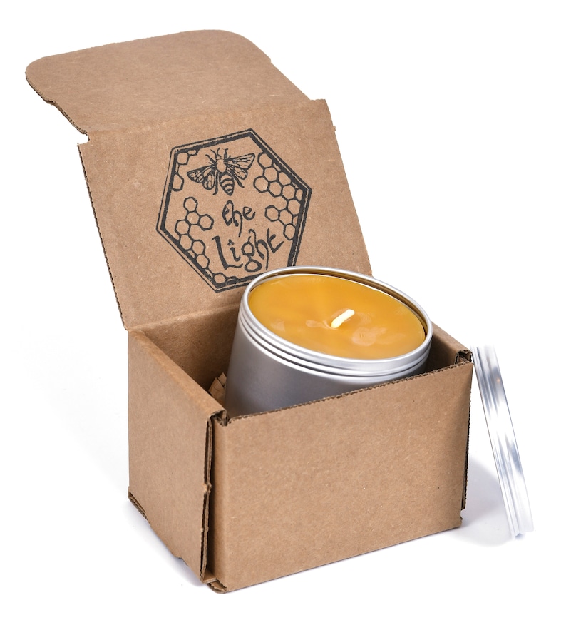 May include: A honey candle in a silver tin with a cardboard box. The box is open and the candle is inside. The box has a logo that says "The Light" with a bee and honeycomb design.