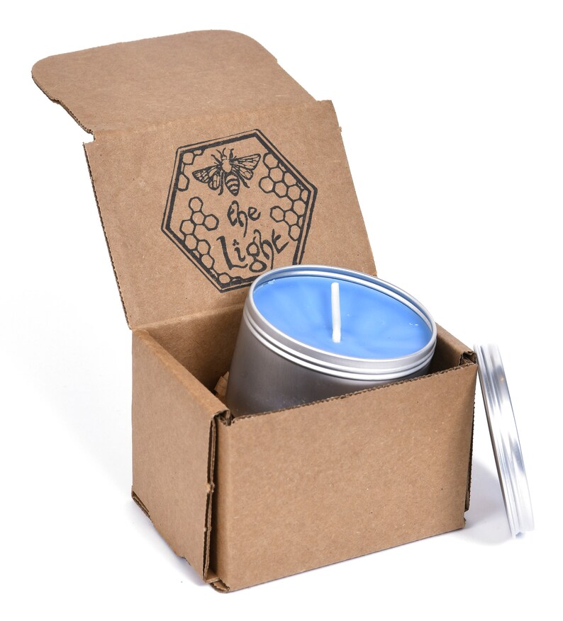 May include: A blue candle in a silver tin with a cardboard box. The box is open and the candle is inside. The box has a logo that says "the light" with a bee and honeycomb design.