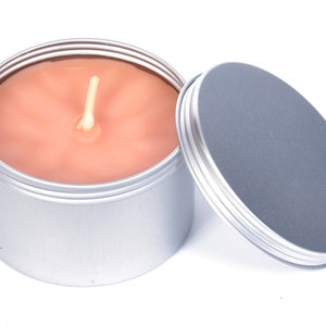 May include: A silver tin candle with a peach-colored wax and a white wick. The lid is off and sitting next to the candle.