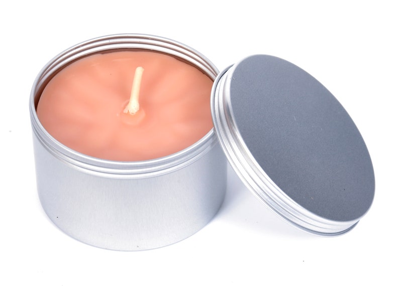 May include: A silver tin candle with a peach-colored wax and a white wick. The lid is off and sitting next to the candle.