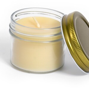 May include: A clear glass jar with a gold lid containing a white candle with a single wick. The lid is off the jar and the candle is lit.
