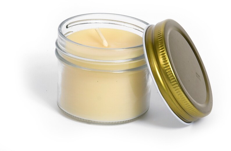 May include: A clear glass jar with a gold lid containing a white candle with a single wick. The lid is off the jar and the candle is lit.