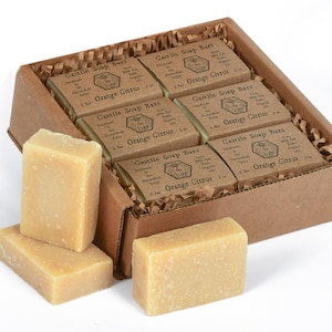 May include: A box of eight bars of orange citrus Castile soap. The soap bars are packaged in a brown cardboard box with shredded paper inside. The soap bars are individually wrapped in paper and have a label that says "Castile Soap Bars" and "Orange Citrus".