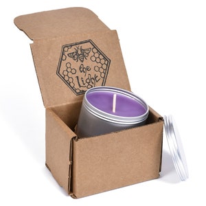 May include: A purple soy candle in a silver tin with a wooden wick. The candle is in a cardboard box with a bee and the text "be the light" printed on the lid.