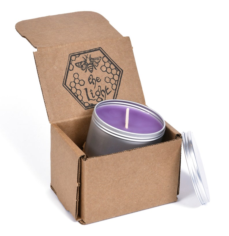 May include: A purple soy candle in a silver tin with a wooden wick. The candle is in a cardboard box with a bee and the text "be the light" printed on the lid.