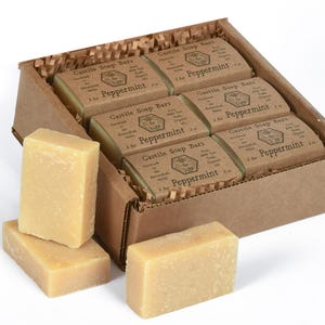 May include: A cardboard box filled with eight bars of peppermint soap. The soap bars are wrapped in brown paper with white text that reads "Castile Soap Bars" and "Peppermint". The box is sitting on a white surface.