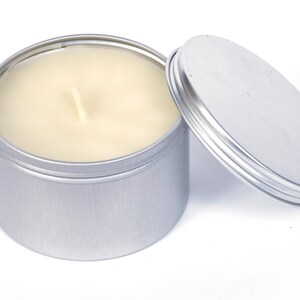 May include: A silver tin candle with a white wax candle inside. The lid is off and sitting next to the candle.