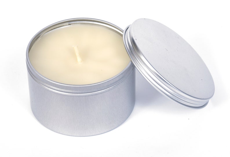 May include: A silver tin candle with a white wax candle inside. The lid is off and sitting next to the candle.