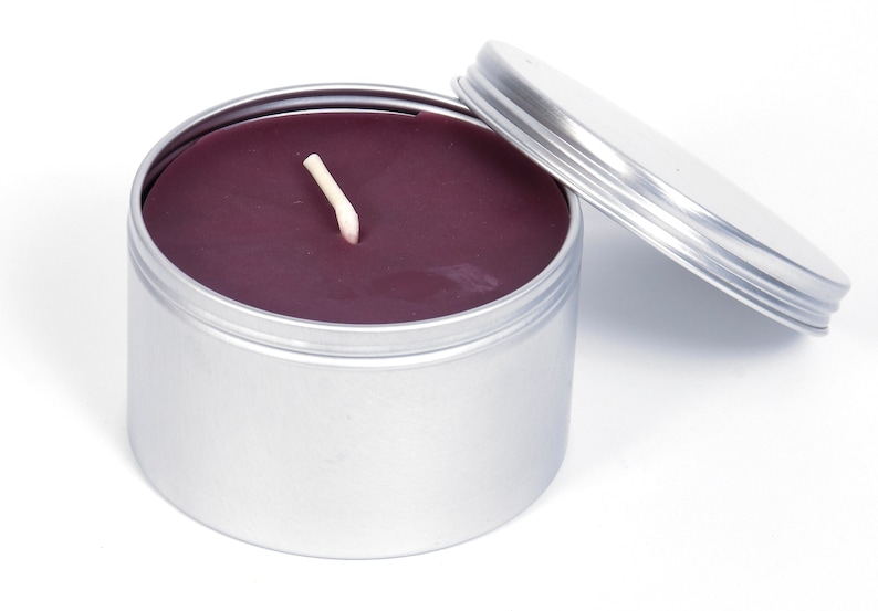 May include: A silver tin candle with a purple wax and a white wick. The lid is off and the candle is sitting on a white surface.