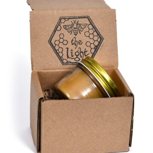 May include: A small glass jar filled with honey, with a gold lid, is sitting inside a cardboard box. The box is open and has a black and white logo on the top that says "the light" with a bee and honeycomb design.