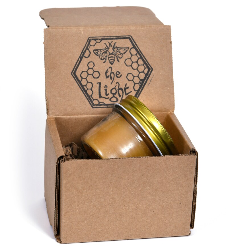 May include: A small glass jar filled with honey, with a gold lid, is sitting inside a cardboard box. The box is open and has a black and white logo on the top that says "the light" with a bee and honeycomb design.