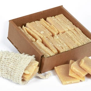 May include: A cardboard box filled with rectangular bars of soap. The soap is a light beige color. A small, beige, woven bag with a few bars of soap inside is in the foreground.