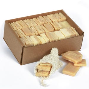 May include: A cardboard box filled with rectangular bars of natural soap. The box is sitting on a white surface. A small burlap bag filled with soap bars is next to the box.