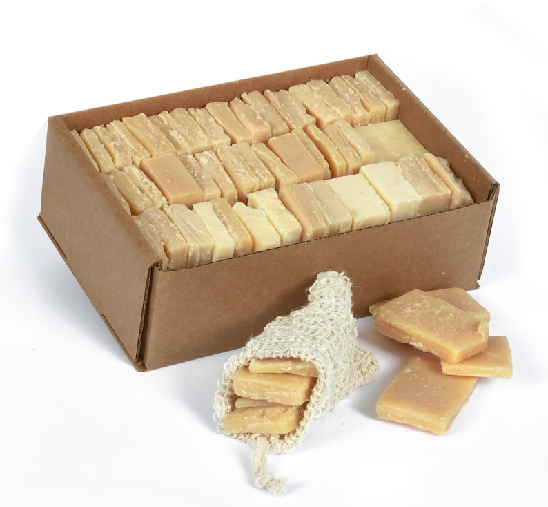 May include: A cardboard box filled with rectangular bars of natural soap. The box is sitting on a white surface. A small burlap bag filled with soap bars is next to the box.