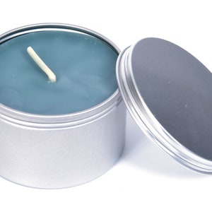 May include: A silver tin candle with a blue wax top and a white wick. The lid is off and sitting next to the candle.