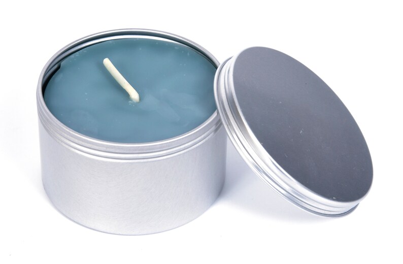 May include: A silver tin candle with a blue wax top and a white wick. The lid is off and sitting next to the candle.