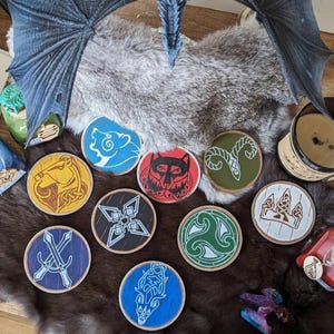 Puede incluir: Un conjunto de posavasos redondos de madera con varios diseños celtas y de animales. Los posavasos tienen un borde de madera natural e imágenes pintadas de colores, incluyendo un oso, un lobo y un carnero. Se muestran sobre una superficie de piel sintética.