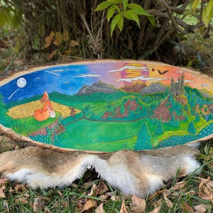 May include: A hand-painted, oval wooden plaque featuring a whimsical landscape scene. The artwork depicts a fox under a moonlit sky, transitioning to a mountain range, castle, and sunset. The plaque is set on a bed of fur and grass.