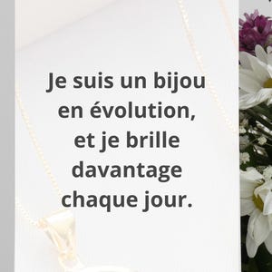 May include: A gold pendant necklace with a small, detailed charm is displayed on a white card. The card has the French text: "Je suis un bijou en évolution, et je brille davantage chaque jour." Flowers are in the background.