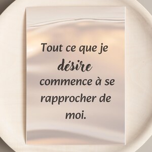 Könnte beinhalten: Eine rechteckige Karte mit dem französischen Text "Tout ce que je désire commence à se rapprocher de moi" in schwarzer Schrift. Der Hintergrund hat einen weichen, verschwommenen, hellfarbigen Farbverlauf.