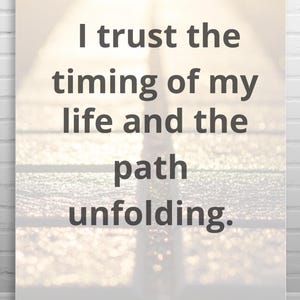 Puede incluir: Un póster motivacional con el texto "I trust the timing of my life and the path unfolding." en gris oscuro sobre un fondo borroso y claro. El póster se muestra en una pared de ladrillo blanco y un suelo de madera.