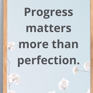 Puede incluir: Impresión enmarcada con el texto "Progress matters more than perfection" en gris oscuro sobre un fondo azul claro. Flores y ramas blancas en las esquinas. El marco es de color marrón claro.