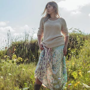 Op de afbeelding: Een vrouw in een veld draagt een crèmekleurige gebreide trui en een rok met bloemenprint. De rok heeft een lichtblauwe basis met een bloemenpatroon in tinten paars, roze en groen. Ze draagt ook bruine laarzen.