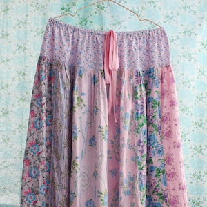 May include: A long, flowing skirt with a patchwork design. The skirt features panels of fabric in various floral patterns, including blue, pink, and lavender. A pink ribbon tie is visible at the waist. The skirt is hanging on a metal hanger.