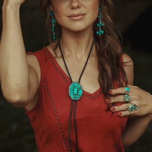May include: A woman wearing a brown cowboy hat, red suede vest, and turquoise jewelry. The jewelry includes earrings, rings, and a bolo tie with a large turquoise pendant. The vest is adorned with metal studs.