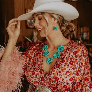 Pode incluir: Uma mulher usando um chap&eacute;u de cowboy branco, um vestido com estampa floral e joias turquesas. O vestido tem um padr&atilde;o floral vermelho e laranja. As joias incluem um colar e brincos com desenhos de flores turquesas. Uma cinta decorativa com uma fivela grande tamb&eacute;m &eacute; vis&iacute;vel.
