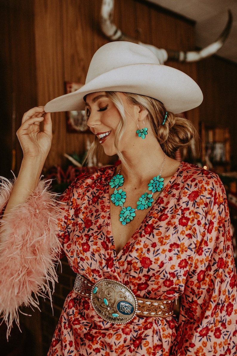 Pode incluir: Uma mulher usando um chap&eacute;u de cowboy branco, um vestido com estampa floral e joias turquesas. O vestido tem um padr&atilde;o floral vermelho e laranja. As joias incluem um colar e brincos com desenhos de flores turquesas. Uma cinta decorativa com uma fivela grande tamb&eacute;m &eacute; vis&iacute;vel.
