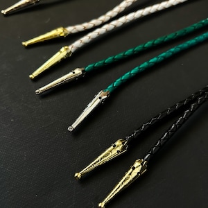 May include: Five different bolo ties with various colored braided cords and silver and gold tips. The bolo ties are arranged in a row on a black surface.