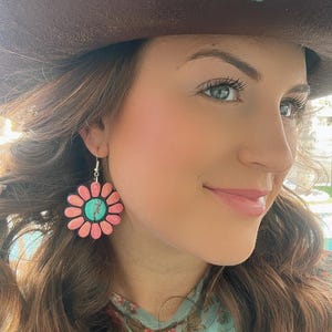 May include: A woman wearing a brown cowboy hat with a turquoise butterfly pin and a floral print shirt. She is wearing pink and turquoise flower earrings.