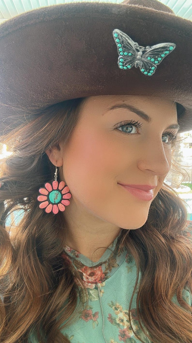 May include: A woman wearing a brown cowboy hat with a turquoise butterfly pin and a floral print shirt. She is wearing pink and turquoise flower earrings.