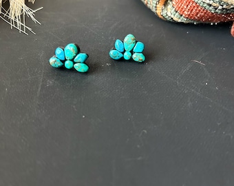 May include: Two turquoise stone stud earrings. Each earring features a cluster of small, irregularly shaped turquoise stones set on a dark base. The earrings are displayed on a dark surface, with a blurred background.