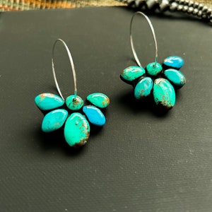 Pode incluir: Um par de brincos de argola de prata e turquesa. Cada brinco apresenta um grupo de pedras turquesas em tons de verde-azulado e azul. As pedras s&atilde;o dispostas em um design semelhante a uma p&eacute;tala e s&atilde;o presas a uma argola de prata.