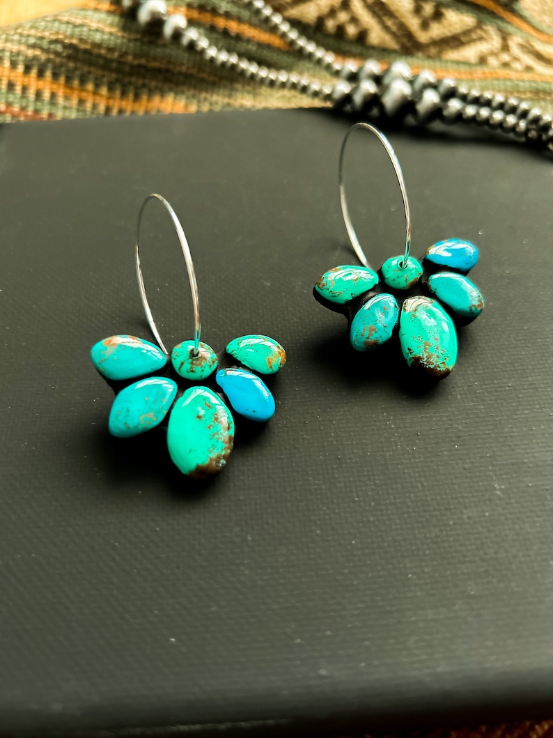 Pode incluir: Um par de brincos de argola de prata e turquesa. Cada brinco apresenta um grupo de pedras turquesas em tons de verde-azulado e azul. As pedras s&atilde;o dispostas em um design semelhante a uma p&eacute;tala e s&atilde;o presas a uma argola de prata.