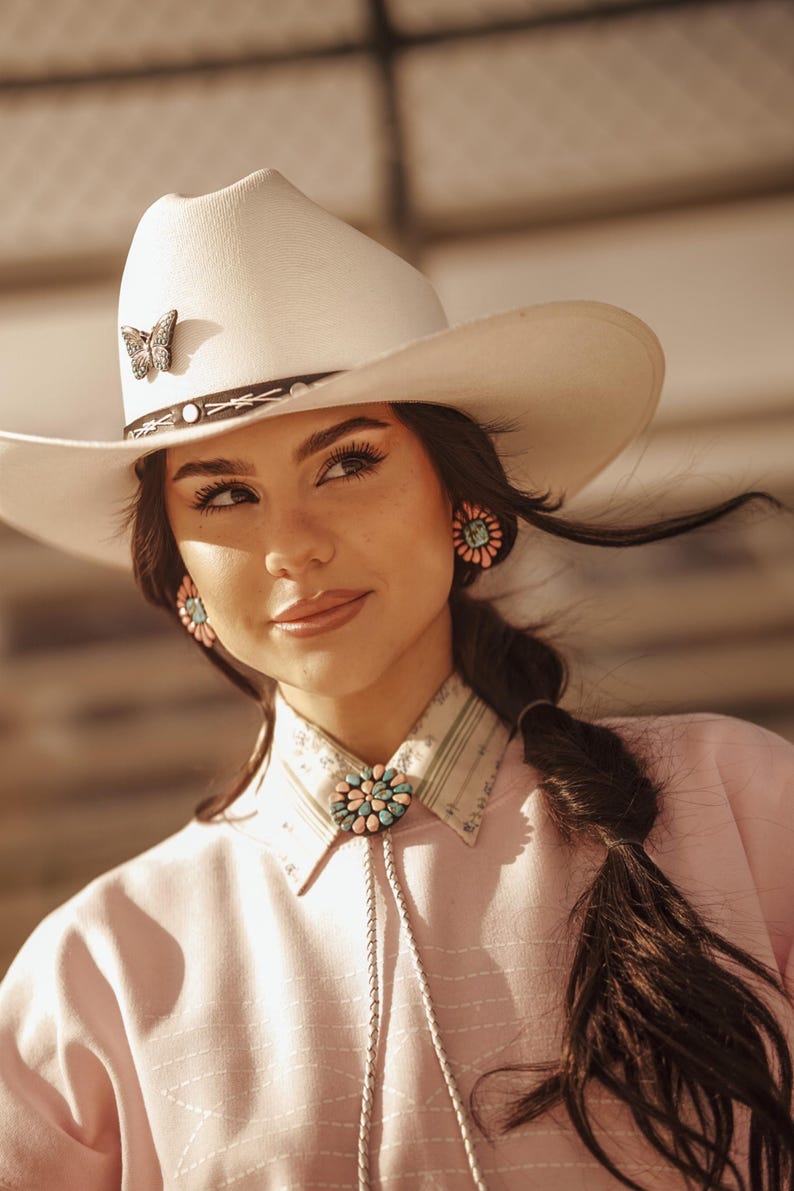 May include: A woman wearing a white cowboy hat with a butterfly pin, a pink shirt with a turquoise and pink bolo tie, and turquoise and pink earrings. Her hair is pulled back in a braid.