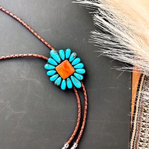 May include: A bolo tie featuring a turquoise and orange flower design. The flower is surrounded by turquoise stones and has an orange center. The braided leather cord is brown, and the tips are silver.