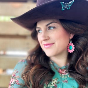 May include: A woman wearing a brown cowboy hat with a turquoise butterfly pin and a floral print shirt with pink flower earrings.