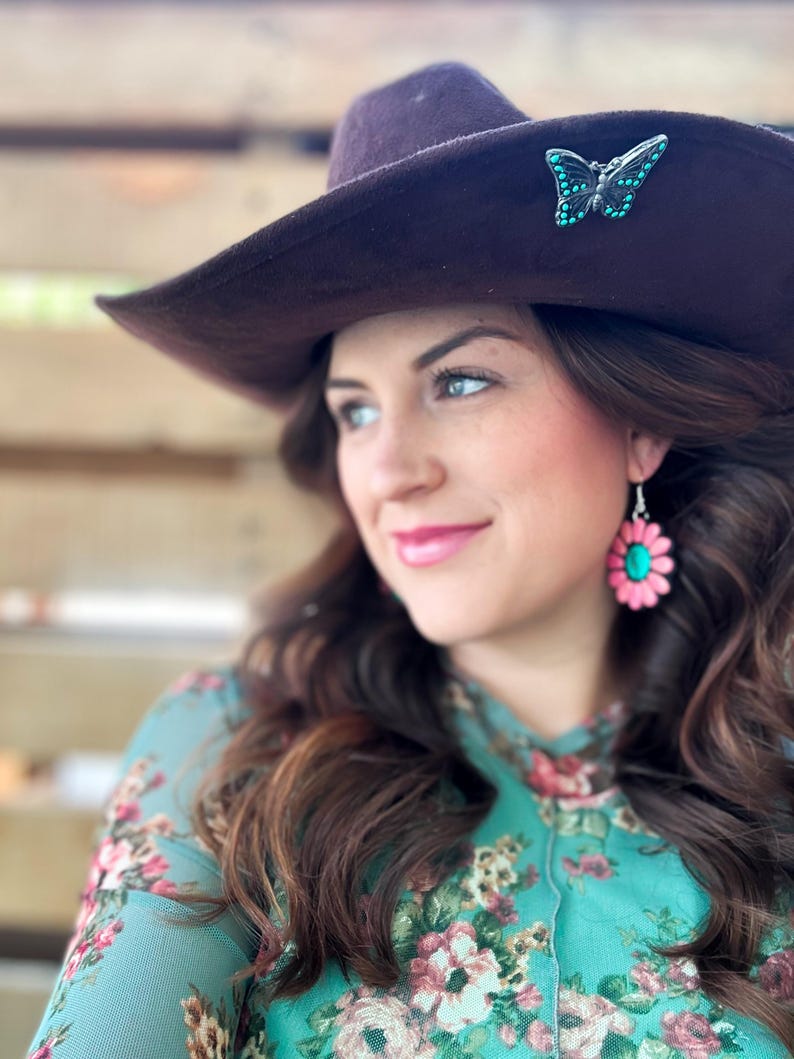 May include: A woman wearing a brown cowboy hat with a turquoise butterfly pin and a floral print shirt with pink flower earrings.