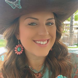 May include: A woman wearing a brown cowboy hat with a turquoise butterfly pin, a floral print shirt, and turquoise and pink flower earrings. She is also wearing a turquoise ring with a flower design.