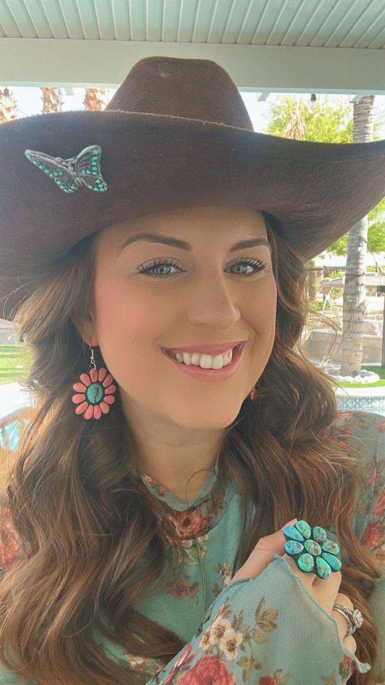 May include: A woman wearing a brown cowboy hat with a turquoise butterfly pin, a floral print shirt, and turquoise and pink flower earrings. She is also wearing a turquoise ring with a flower design.