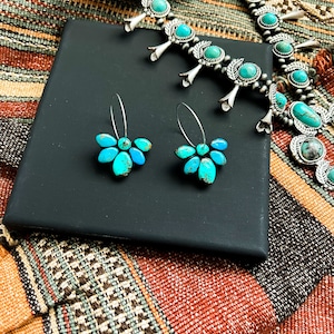 Pode incluir: Um conjunto de colar e brincos turquesa e prata. O colar apresenta pedras turquesas cravejadas em prata, com um par de brincos de flores turquesas a condizer. As joias s&atilde;o exibidas numa caixa preta, sobre um tecido estampado.