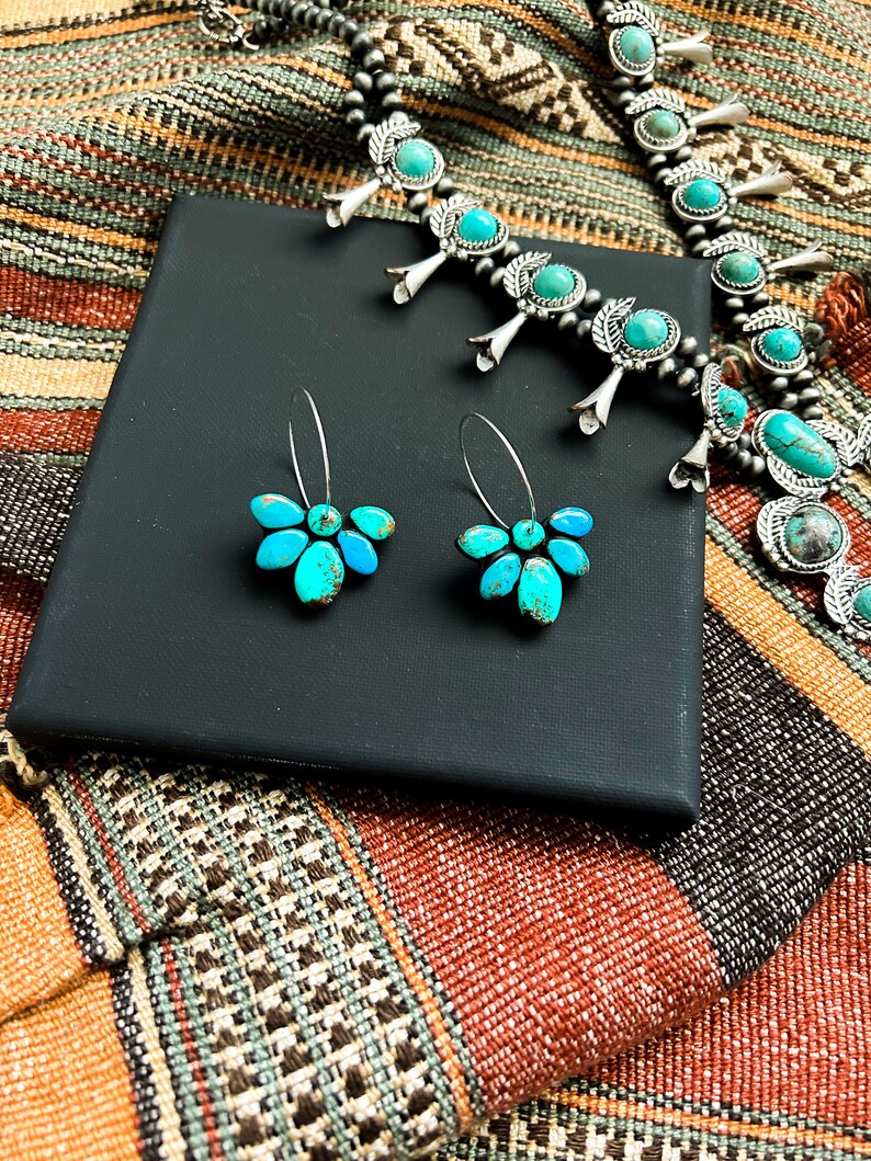 Pode incluir: Um conjunto de colar e brincos turquesa e prata. O colar apresenta pedras turquesas cravejadas em prata, com um par de brincos de flores turquesas a condizer. As joias s&atilde;o exibidas numa caixa preta, sobre um tecido estampado.